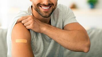 Population inoculation concept. Close up portrait of positive young vaccinated man demonstrating his shoulder with medical plaster after vaccine injection, smiling to camera at home photo