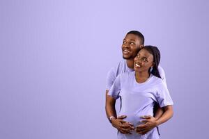 Smiling black husband hugging pregnant wife from back, cheerful couple standing on purple violet studio background, thinking about baby's name or gender looking up at empty speech bubble, free space photo