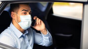 Closeup of millennial arab businessman in protective face mask sitting in car, having conversation on cellphone, shot from automobile window, side view, copy space. Business while COVID-19 pandemic photo