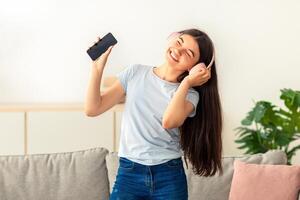 Happy Indian teenage girl with headphones and mobile device dancing to music at home. Joyful adolescent moving to popular song, having great time indoors during coronavirus lockdown photo
