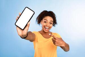 Check this out, cellphone display mockup. Excited African American woman pointing at smartphone with empty screen on blue background, selective focus. Space for mobile app, website or online ad photo