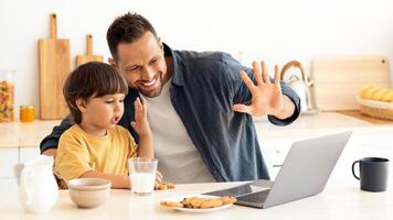 Online communication. Little boy and his daddy calling to relatives via laptop, waving hands to computer and smiling, sitting together at kitchen, free space photo