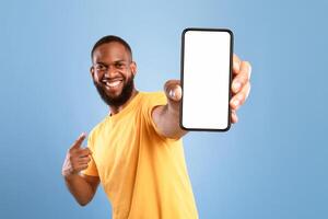 Excited African American guy in casual wear pointing at cellphone with empty screen on blue studio background, space for website or mobile app design. Cellphone display mockup. Selective focus photo