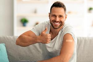 Healthcare and immunization concept. Confident happy guy approving vaccinations, showing medical plaster on shoulder after injection and gesturing thumb up, smiling to camera on couch, free space photo