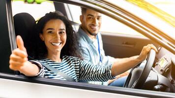 Cheerful arab man instructor examinating happy lady student, taking notes at chart while sitting by smiling young woman driving auto and showing thumb up, side view, copy space, sun flare photo