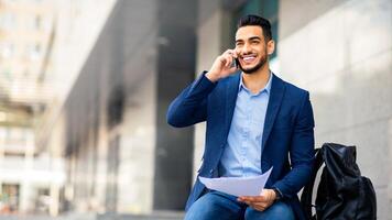 Cheerful arab guy in formal suit sitting on the street by office building, holding papers and having phone call after job interview, sharing good news with girlfriend or friends, copy space photo
