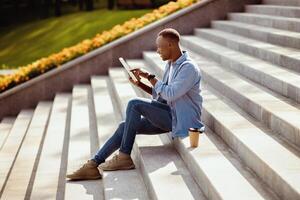 Side view of happy black guy using tablet computer on stone stairs at city park photo