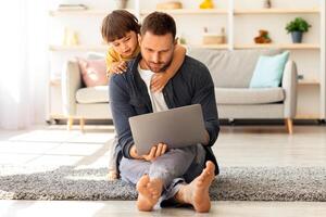 Interest for modern technologies. Young busy man working on laptop from home, his cute little son hugging him and watching at gadget, sitting together on floor at home, free space photo