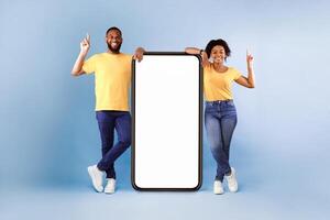 Cheerful African American couple standing next to big smartphone with blank white screen, pointing upwards, recommending new mobile application on blue studio background photo