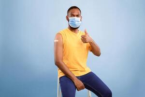 Joyful vaccinated African American guy with adhesive plaster bandage on his arm after flu antiviral vaccine injection showing thumb up gesture on blue studio background photo