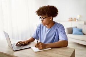 E-learning concept. Smart African American teenager writing in notebook during web lesson at home. Busy black adolescent participating in online lecture or speaking to tutor on internet photo