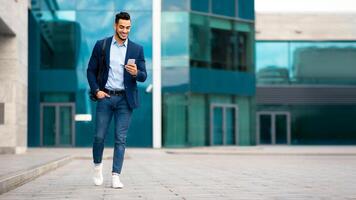 Confident arabic guy programmer with backpack walking by street, checking his mobile phone, chatting with friends, going home from office, panorama with copy space, full length photo