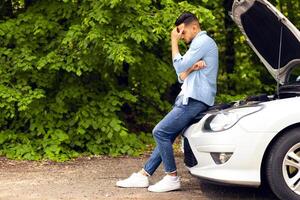 Puzzled arab man standing by open car hood, looking down, having automobile crush in the middle of the road. Nervous middle-eastern guy driver waiting for car service, panorama with copy space photo