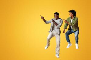 Look At This. Two Joyful Black Guys Pointing Aside While Jumping In Air Over Beige Studio Background, Cheerful African American Friends Having Fun And Demonstrating Free Copy Space, Panorama photo