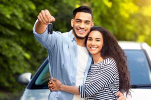 Happy young middle-eastern couple showing brand new car key, man and woman hugging and smiling, standing by new luxury auto, copy space. Car renting, leasing, buying, auto retail concept photo