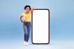 Happy African American woman leaning on big smartphone with blank white screen, smiling at camera on blue studio background, mockup for mobile app, website, your advertisement design photo