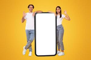 Excited Casual Couple Leaning On Big Smartphone With White Empty Screen Showing Okay Sign Gesture, Cheerful Guy And Lady Recommending New App Or Website, Standing At Orange Wall, Mock Up, Full Body photo
