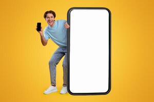 Full Body Length Of Cheerful Man Peeking Out Standing Behind Big Smartphone With White Blank Screen, Excited Guy Holding Cell Phone Presenting New App Showing Copy Space For Website Design Mock Up photo