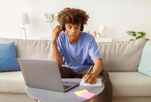 Smart African American teenager attending online lesson or webinar, sitting on couch, using wireless headset and laptop, taking notes indoors. Remote education, home schooling concept photo