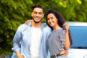 Happy young middle-eastern couple showing key from new car, smiling arab man and woman hugging, standing by new luxury auto, copy space. Car renting, leasing, buying, dealership, auto retail concept photo