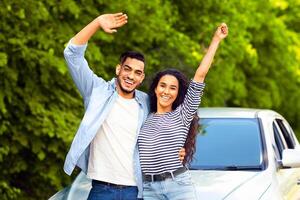 Emotional multiracial couple standing by nice auto at countryside, embracing and waving at camera, copy space. Happy middle-eastern man and woman having car trip together, renting automobile photo
