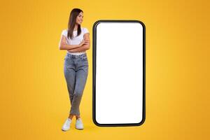 Cheerful smiling casual lady standing with crossed hands near giant big cell phone, empty white screen template, orange studio wall. Mock up for app or website, free copy space, full body length photo