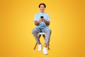 People And Technology Concept. Portrait of smiling young man using smartphone sitting on chair isolated on orange studio background, looking at camera. Guy chatting online, browsing social media photo
