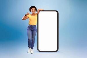 Mobile offer. Happy African American lady leaning on huge cellphone with empty white screen, showing thumb up, recommending cool new app or website, offering space for your ad, mockup photo