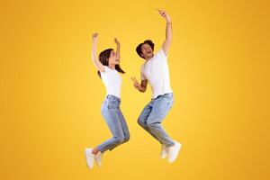 Full body length shot of happy excited couple taking selfie, using smartphone, jumping high up in the air, isolated over orange yellow studio background, vertical banner, showing peace sign gesture photo
