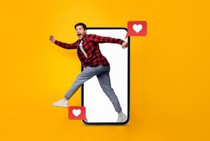 A man joyfully runs out of a smartphone, engaging with social media notifications symbolizing love. The bright yellow background enhances the theme of connection and modern technology. photo