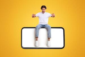 Mobile App Advertisement. Handsome Excited Man Sitting On White Empty Smartphone Screen And Pointing Fingers Down Isolated Over Orange Studio Background. Check This Out, Cell Phone Display Mock Up photo