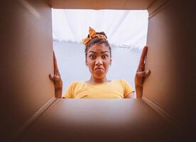 Bad Delivery Service Concept. Angry dissatisfied black woman unpacking and looking in cardboard box, received damaged item, feeling upset with wrong parcel, view from the inside of package photo