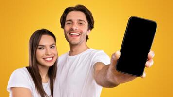 Handsome guy holding and showing smart phone with black empty screen in hand close up to camera, mock up macro, young lady embracing man, family recommending app on mobile phone, selective focus photo