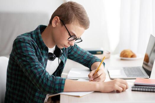 Serious schooler doing homework at his room, writing in notebook, using laptop, side view, empty space photo