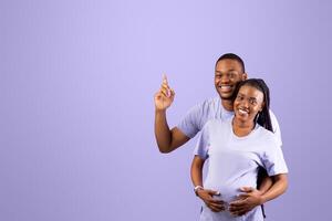 Portrait of smiling African American family hugging, man embracing his pregnant wife and pointing finger up at blank speech bubble cloud, thinking about baby's gender, choosing name. Free copy place photo
