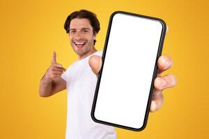 Mobile App Advertisement. Handsome Excited Man Showing Pointing At White Empty Smartphone Screen Posing Over Orange Studio Background, Smiling To Camera. Check This Out, Cellphone Display Mock Up photo