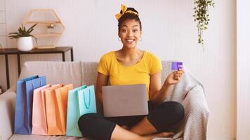 Online Shopping Concept. Excited overjoyed black woman showing credit card and using laptop, ordering clothes and purchases via internet, sitting on the couch with colorful bags in living room at home photo