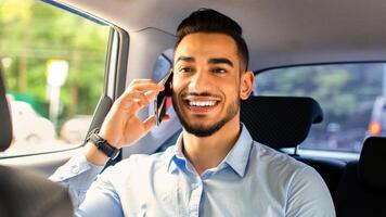 Cheerful middle-eastern young man manager talking to business partner on cellphone while going to office by car, sitting at car back seat, closeup portrait. Businessman lifestyle concept photo