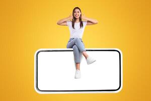 Excited Woman Sitting On Big Smartphone With Blank White Screen And Gesturing Thumb Up Sign, Cheerful Lady Recommending New App Or Website, Isolated On Yellow Background, Mock Up, Creative Collage photo