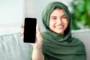 Muslim teenage girl in hijab showing cellphone with blank screen at home, mockup for mobile app or website design. Happy adolescent in headscarf holding smartphone with empty space. Selective focus photo