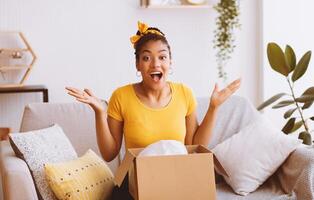 Wow, It's Amazing. Portrait of excited emotional african american woman received package with gift present, sitting with carton cardboard box on the couch in living room at her home, spreading hands photo