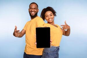 Cheerful African American couple holding tablet pc together, pointing at empty screen with space for mockup, promoting new application, website, advertising your product, blue background. photo