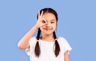 Korean Little Girl Looking Through Okay Sign Covering Eye Posing On Purple Studio Background. Everything Is OK. Free Space photo
