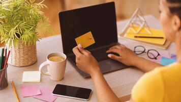 Online Shopping Concept. Over the shoulder view of african american woman holding credit card in hand and using laptop with black blank screen, typing on keyboard, copy space, phone on the table photo