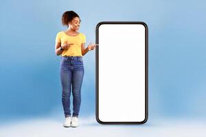 Cheerful African American woman standing next to huge smartphone, pointing at empty screen, advertising new mobile application or website. Blue studio background, cellphone display mockup photo