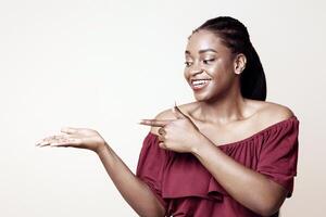 Pretty millennial black lady in stylish casual outfit pointing at something invisible on her palm and smiling, holding something, posing on beige studio background, panorama with copy space photo