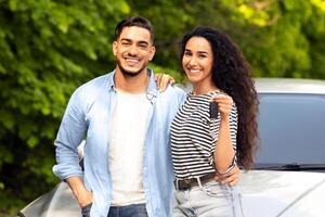 Cheerful beautiful middle-eastern couple young man and woman standing outside by nice auto, hugging and showing key from automobile, having car trip together. Car renting, leasing, sharing concept photo