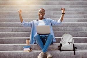 Excited African American student gesturing YES in front of laptop computer on stairs downtown photo