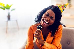 Birthday Fun. African American Woman Singing Song With Eyes Closed Holding Microphone Having Karaoke Party Sitting In Chair At Home, Wearing Festive Hat. B-Day Leisure Concept photo