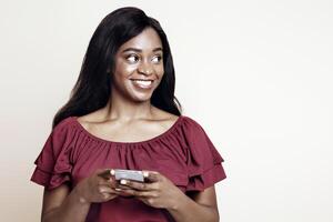 Pretty long-haired young black woman with newest cellphone looking at copy space over beige studio background, using nice dating, entertaining mobile application on smartphone, panorama photo